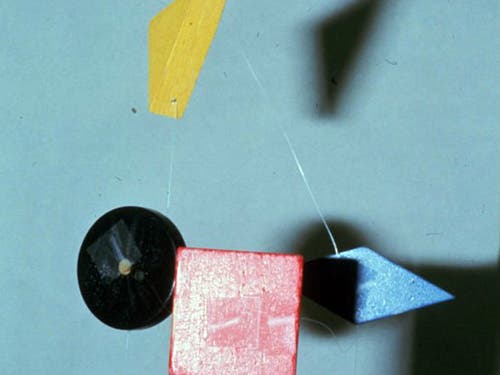 A photo of a mobile of hanging blocks: a yellow triangle at the top, and a black disc, a red square, and a blue diamond hang below. 