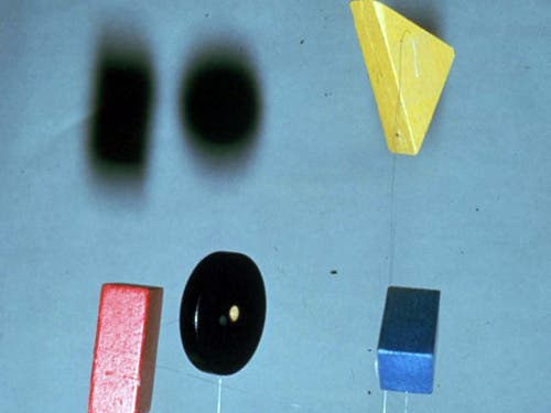 A photo of a mobile of wooden blocks: A red rectangle, a black disc, a blue rectangle, and a yellow triangle. 