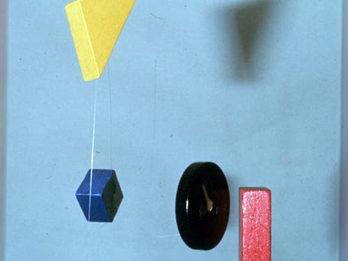 A photo of a mobile with four wooden blocks: a yellow triangle, a blue cube, a black circle, and a red square. 