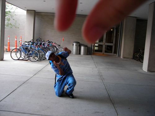 A trick photo where two fingers are in the foreground close to the camera, and a person is kneeling on the ground int he background. The image looks as if two giant fingers are about to pick up the person. 