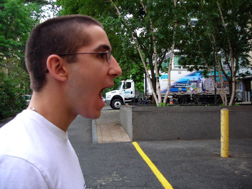 A trick photo where a person stands in profile in the foreground with their mouth open. A parked truck is in the background. It looks as if the person is about to eat the truck. 