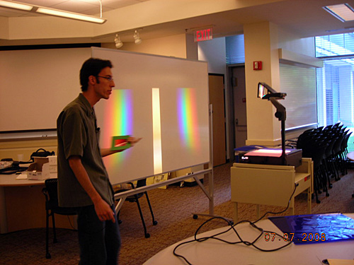 The instructor stands at the front of a classroom. The whiteboard next to him has two rainbows of light projected onto it. 