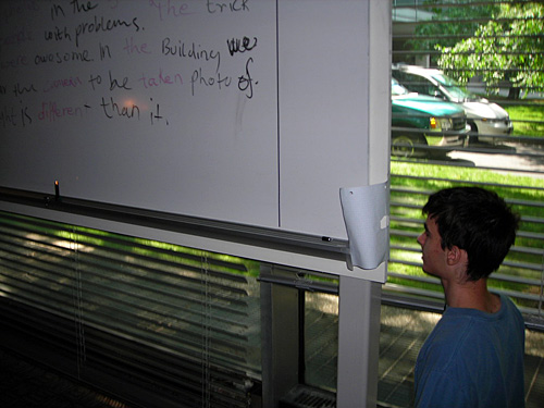 A photo of a student kneeling at the side of a whiteboard looking through a hole made in a white piece of paper.