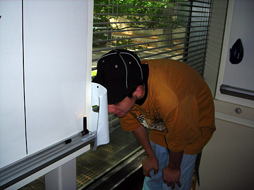 A student bends down at the side of a whiteboard and peers through a hole made in a piece of white paper. 