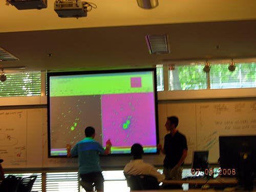 An instructor and a student stand in front of a screen at the head of the class. The screen is filled with a green rectangle, a black square, and a pink square. 