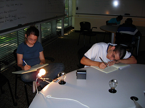 Two students take notes at a table while looking at a glowing lightbulb. 