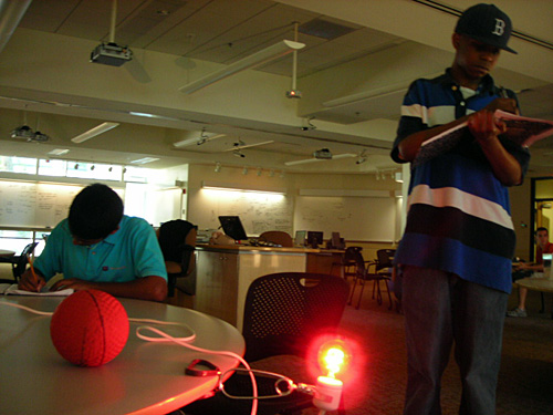 Two students write in notebooks as they look at a ball and a red bulb. 