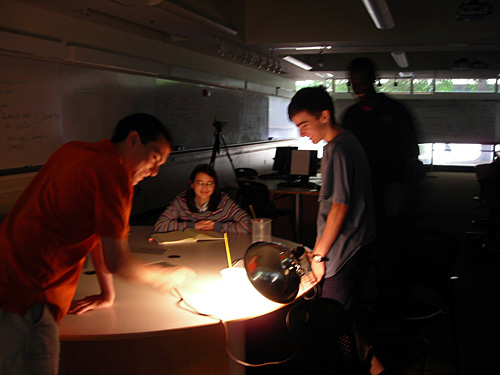 Students stand at a round table in a darkened room observing a thermometer in a beaker of water, while a bright light shines on it. 