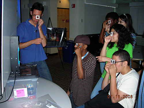 A photo of a demonstration of diffraction emission. A group of people al hold up small squares to their eyes as they view a light source. 