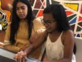 Two girls sitting in front of a laptop, with one typing on laptop and the other girl looking at the laptop screen