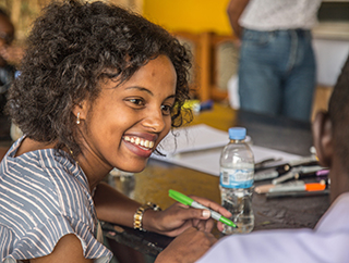 A young African woman is smiling as she workshops ideas with other local entrepreneurs at a large table. 