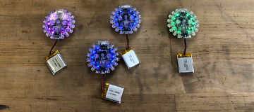 Four Bluefruit boards attached to batteries and lit with different colors.