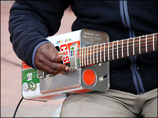 A picture of a man playing an electric guitar. The guitar’s body is made out of an old Castrol oil can, which has been painted with South African colors (black, green, and red).