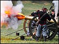 A photograph of a group of people, dressed in Civil War uniforms, standing by a cannon. The cannon has just gone off.