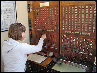 A photograph of a woman plugging a cable into a slot on an old telephone switchboard.