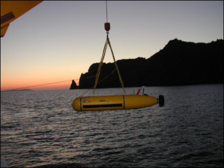 A small submarine-type vehicle being lowered into the water from a large ship at sunset near a rocky coast.