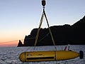 A small submarine-type vehicle being lowered into the water from a large ship at sunset near a rocky coast.