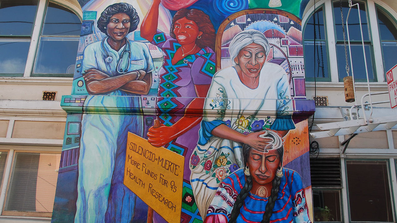 A mural on the side of a building featuring six women dressed in different outfits, including indigenous dress and a woman in medical scrubs. A sign reads "Silencio=Muerto More funding for Women's Health Research"