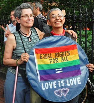 A middle-aged, grey-haired female couple pose with their arms around each other,holding a sign with the words: Equal Rights For All. Love Is Love.
