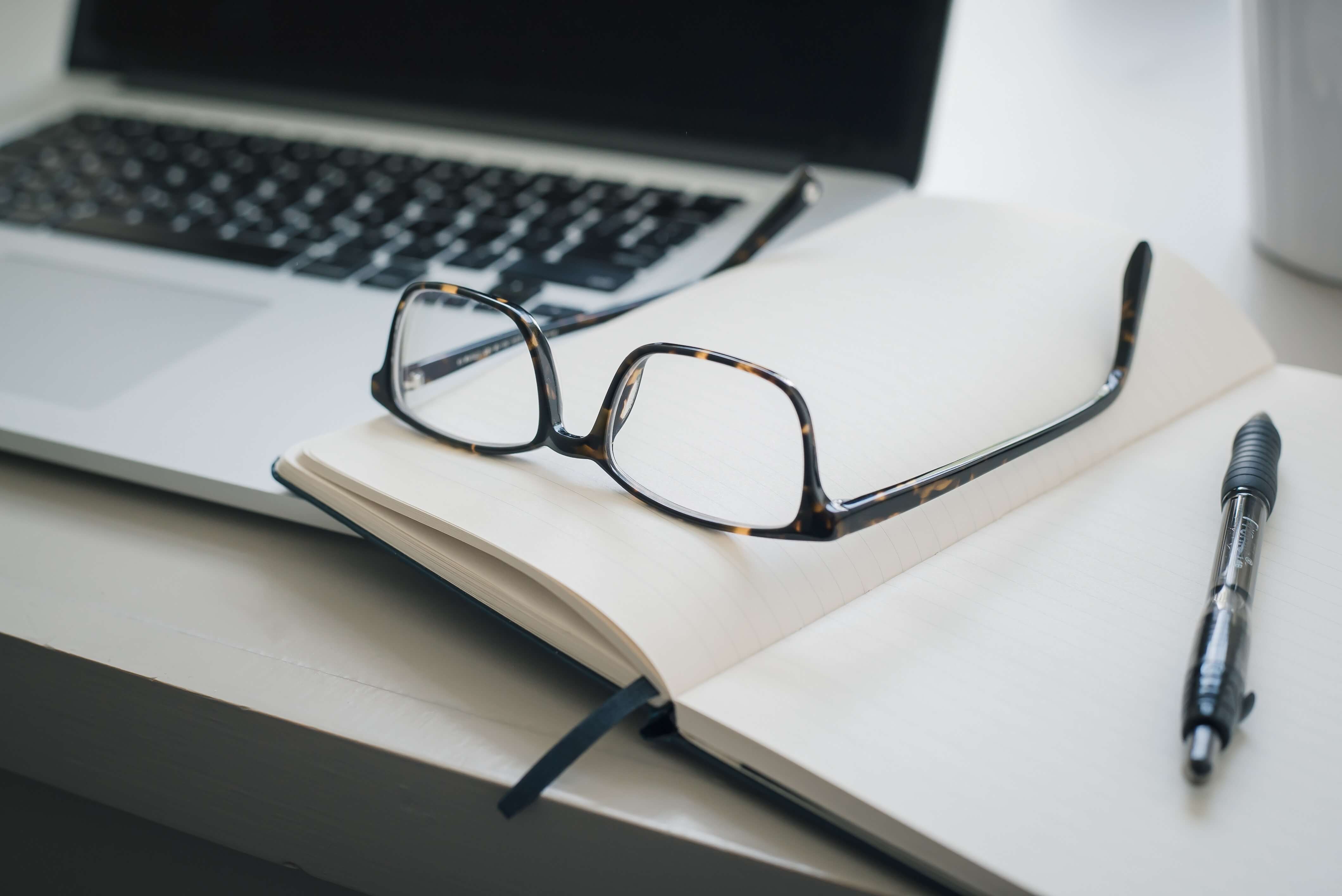 A photo of a laptop, a pair of glasses, and an open notebook with a pen resting on it. 